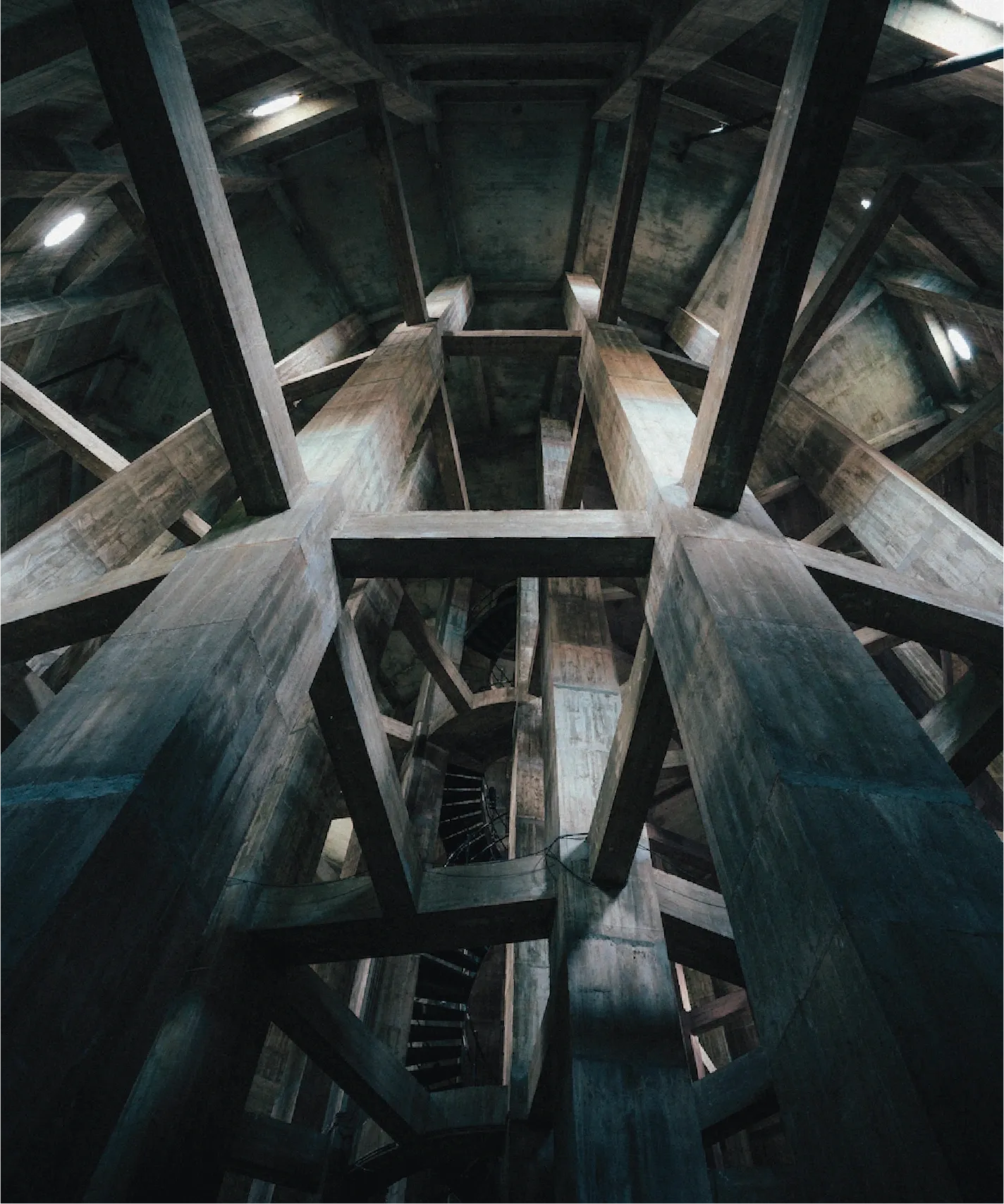Looking up into the dim interior of Brønshøj Water Tower — a lattice of raw concrete columns and beams, faint daylight through slit windows.