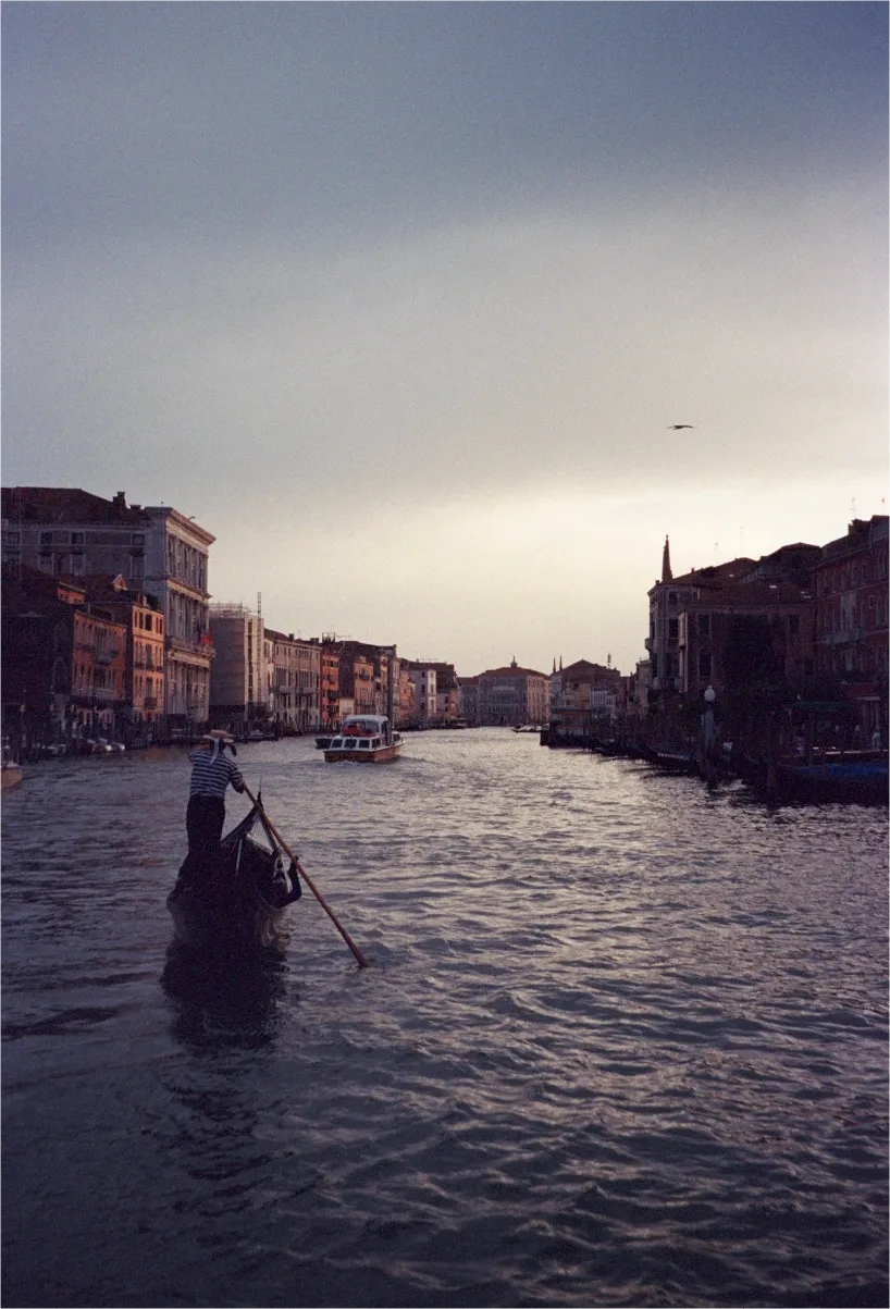 A gondolier poling along Venice's Grand Canal at dusk, buildings lining the water on both sides.
