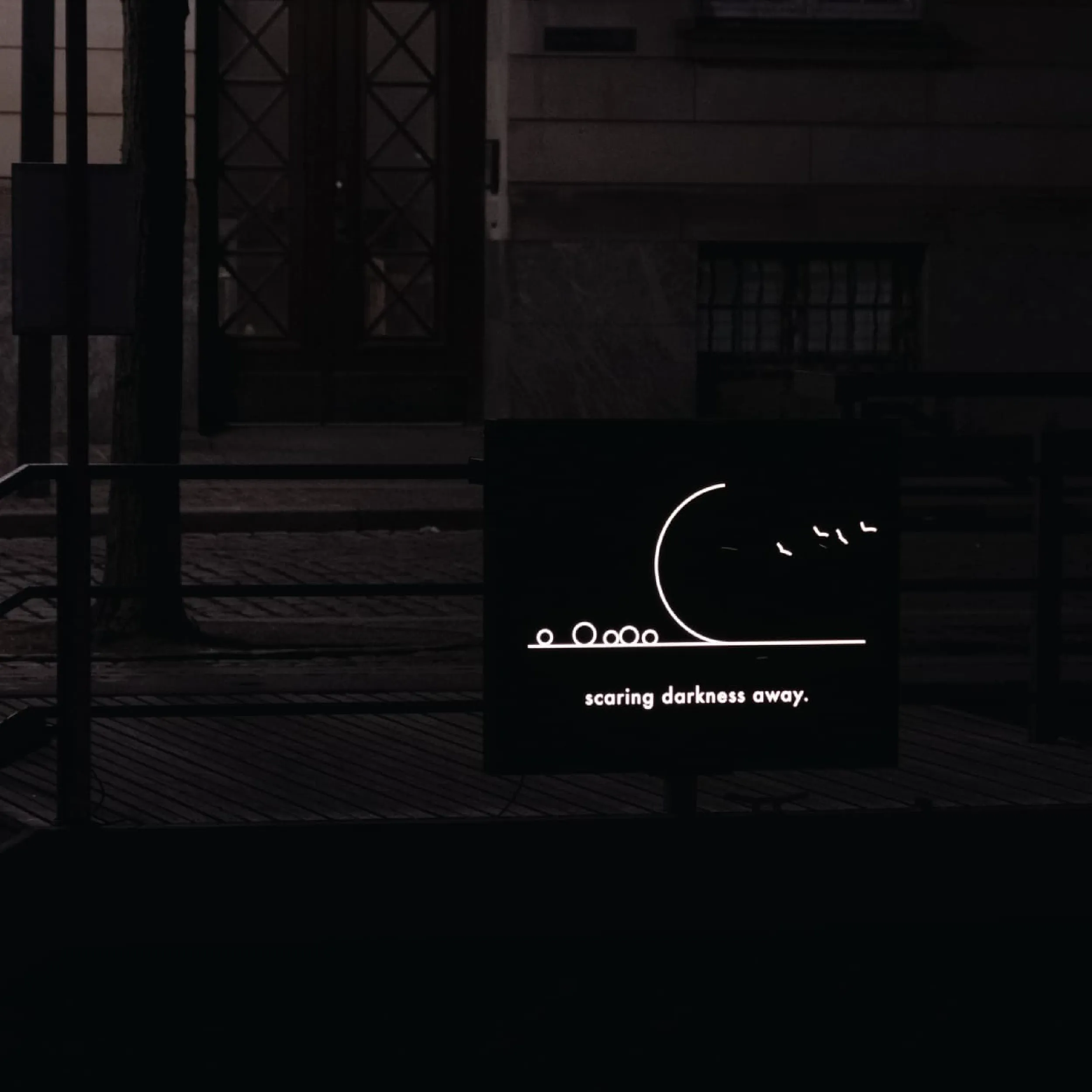 An illuminated sign showing a line-drawn moon, birds and circles, installed on a dark street in Copenhagen at night.