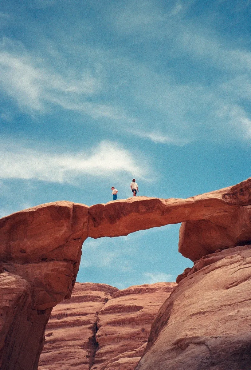 Two people standing on top of a natural rock bridge in the desert, sky visible through the arch below.