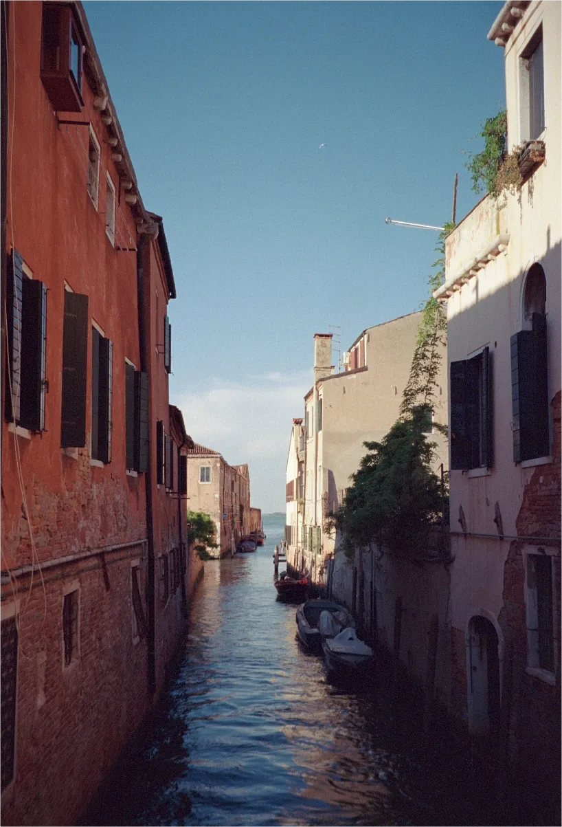A narrow Venice canal lined with weathered orange and cream buildings, boats moored along the water.