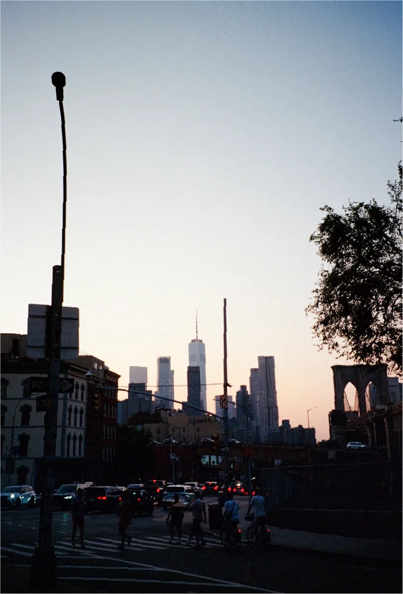 A dusk street scene near the Brooklyn Bridge, traffic and pedestrians in silhouette against the Manhattan skyline.