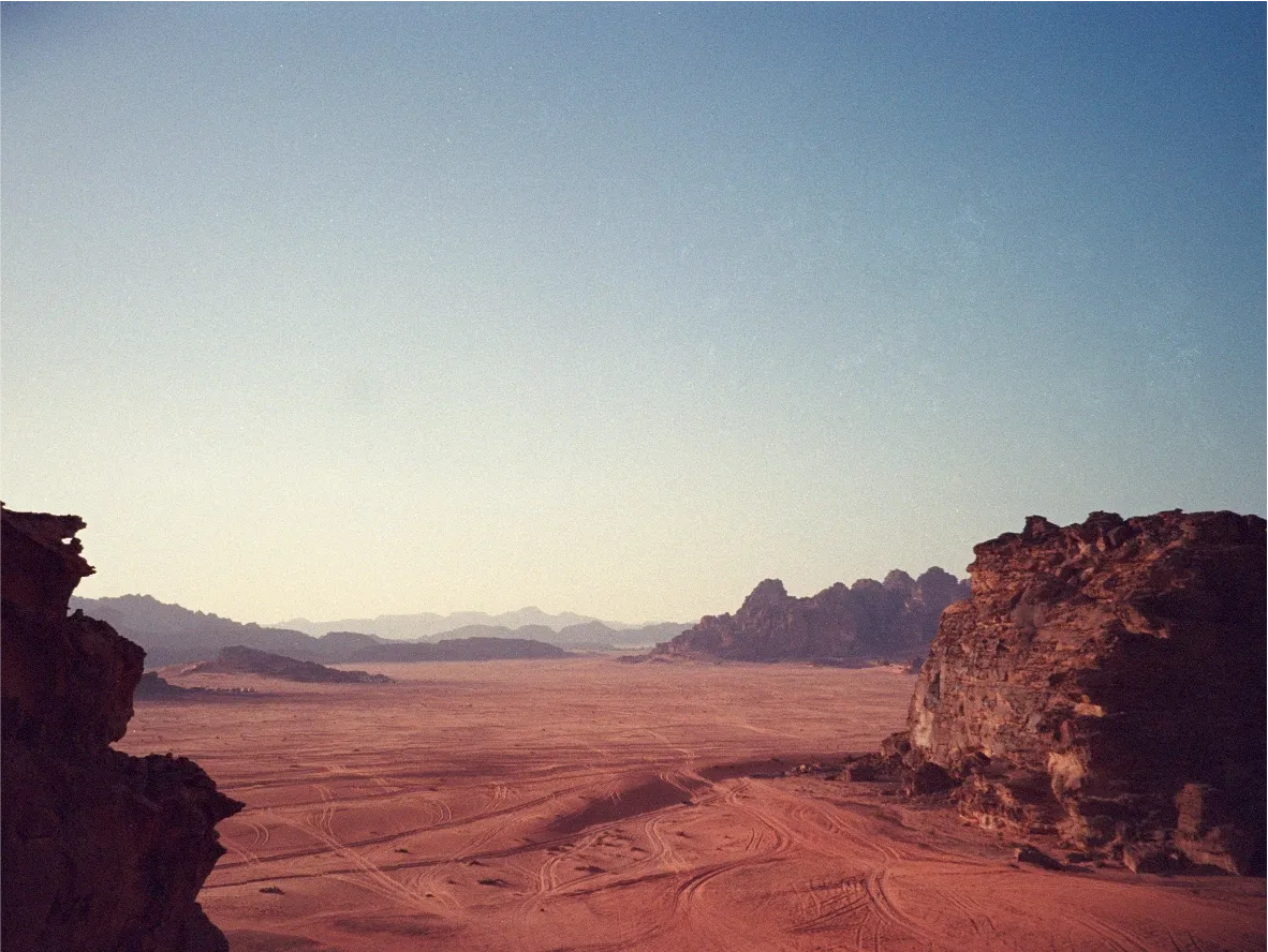 A wide desert valley seen from a clifftop, red sand and distant rock formations under a hazy sky.
