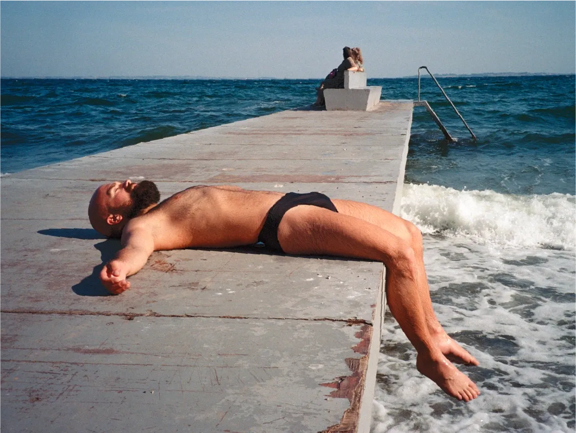 A person sunbathing on a wooden swim pier, legs dangling into the waves below.