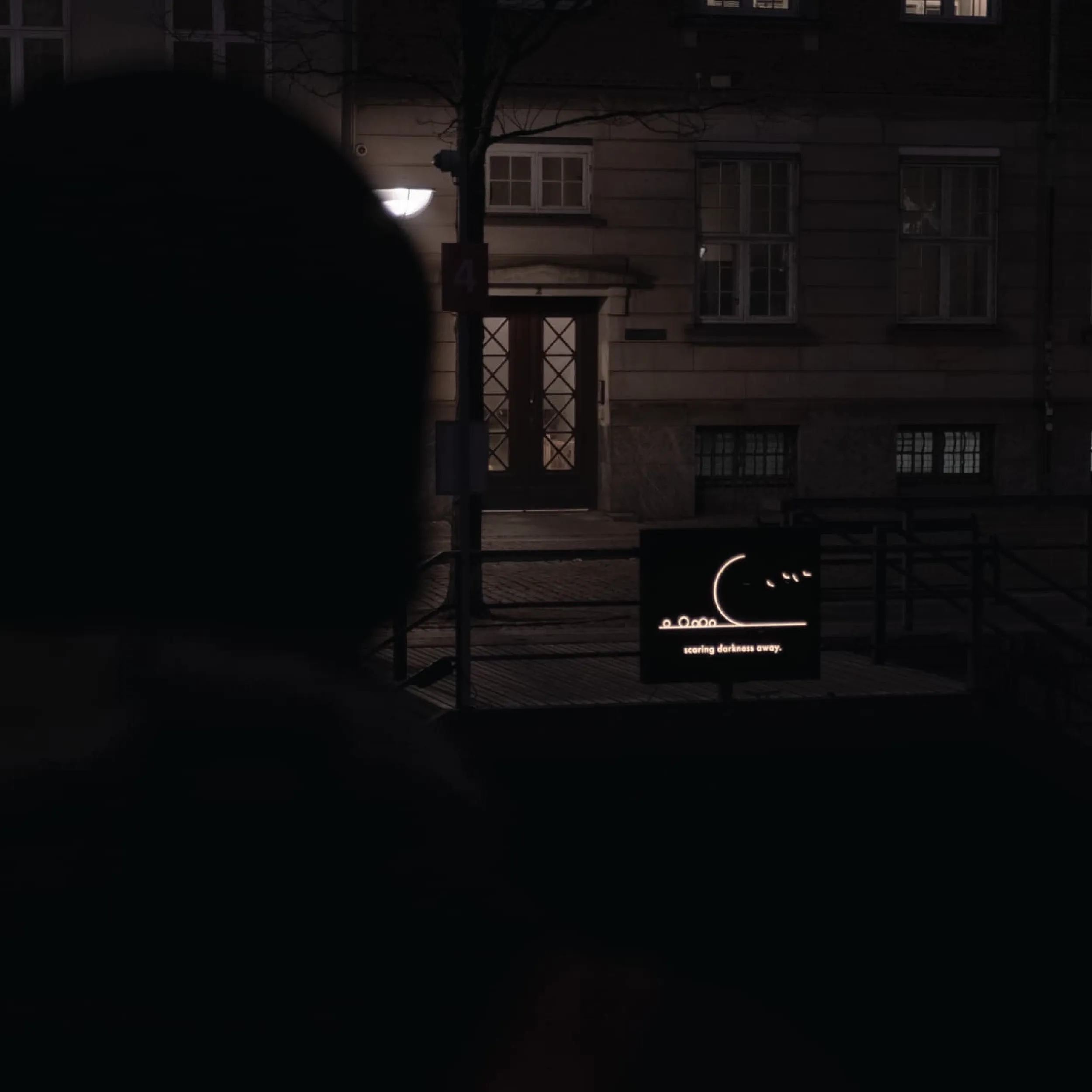 A person's silhouette in the foreground, with the illuminated installation visible on a building facade across the street at night.