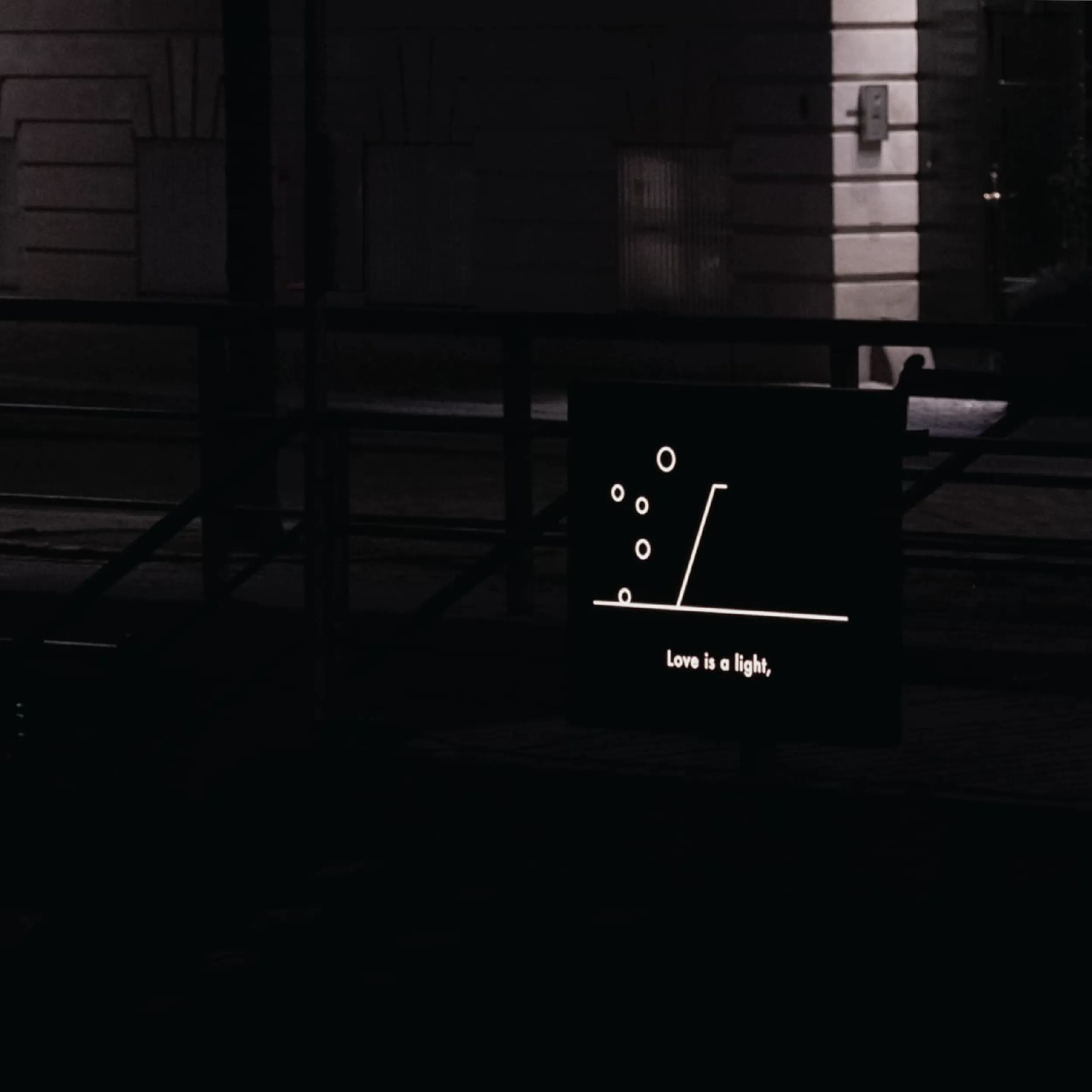 Close-up of the illuminated sign at night, showing the line-drawn rising mark and circles, captioned 'Love is a light,'.