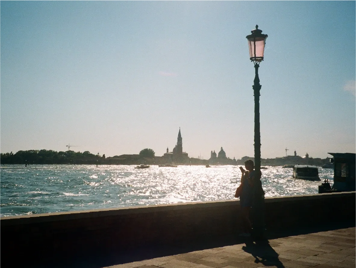 A person silhouetted beside a lamppost on the Venice waterfront, sunlit lagoon and a bell tower in the distance.