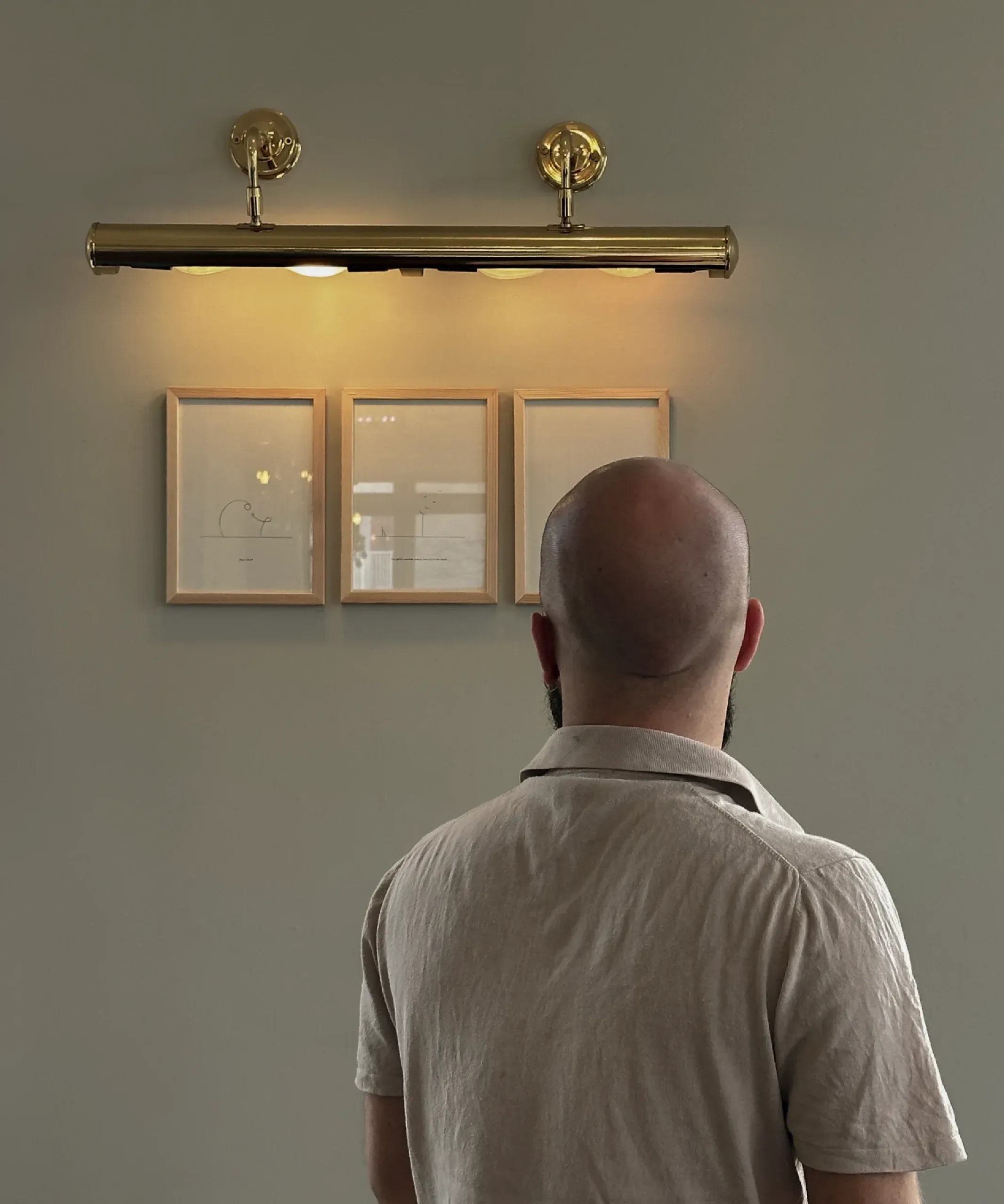 Frederik Smal, seen from behind, looking up at his framed drawings lit by a picture light on a gallery wall.