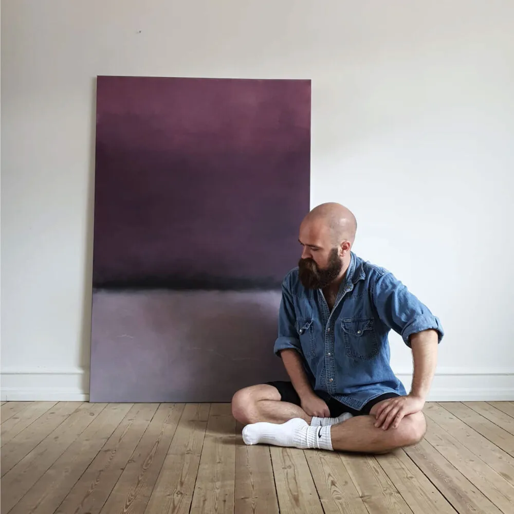 Frederik Smal seated cross-legged on a wooden floor beside a large oil painting in gradient purple and grey tones.